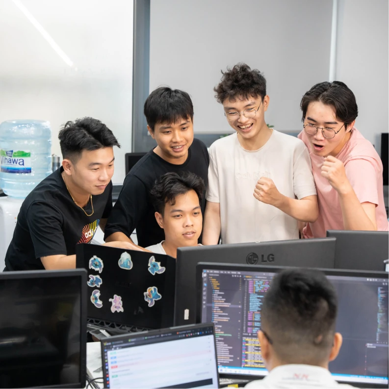 Đội lập trình MONA đứng chụm lại xem một màn hình code trong văn phòng