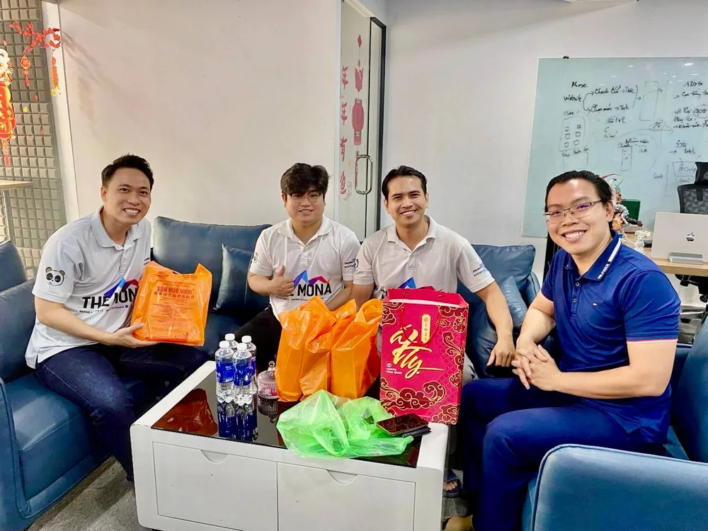 Ba nhân sự MONA mặc áo đồng phục ngồi trên ghế sofa tiếp khách tại văn phòng, trên bàn có quà Tết